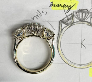 three stone diamond engagement ring sitting side on next to the designer's drawing