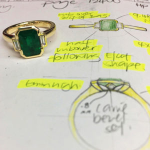 custom made emerald ring with baguette diamond shoulder sitting on the original design drawing